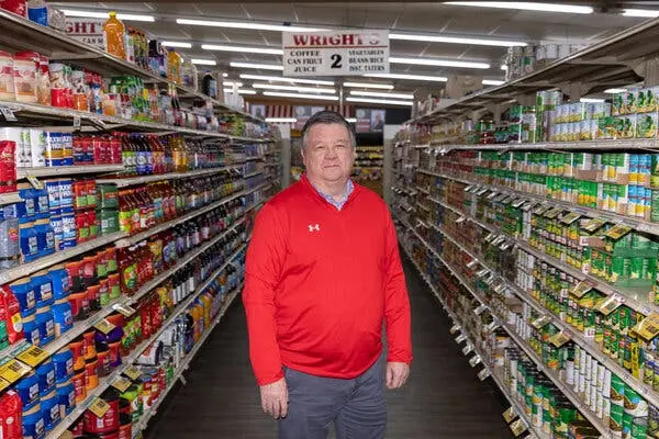 جیمی رایت، با پیراهن قرمز، در یکی از راهروهای فروشگاه‌ خود ایستاده است.