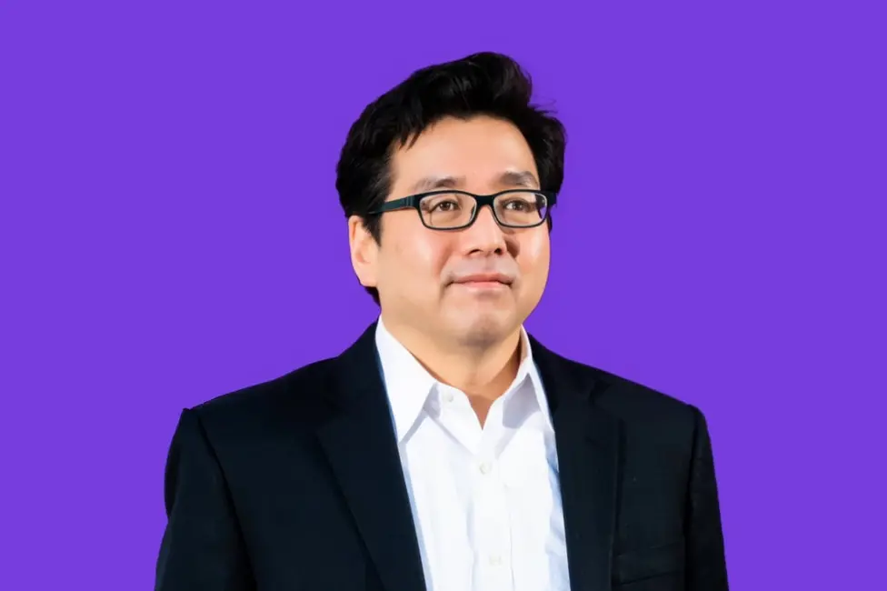 مردی با موهای مشکی و عینک، پیراهن سفید و کتوشلوار تیره بر شلوار بژ پوشیده است.