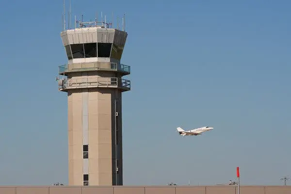 برج کنترل یک فرودگاه با سقف شیشهای و آنتنها؛ هواپیمای سفید در برابر آسمان آبی روشن به پرواز در میآید.