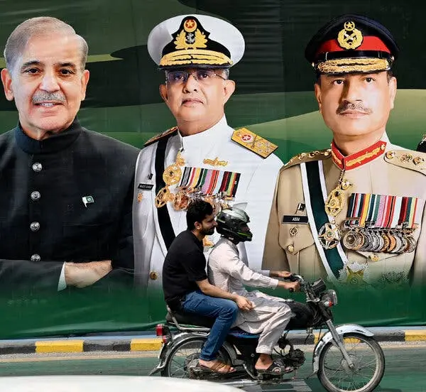 یک موتورسیکلت که از پشت یک پوستر بزرگ عبور میکند؛ این پوستر چهار مرد را نشان میدهد که سهتای آنها یونیفرم نظامی پوشیدهاند