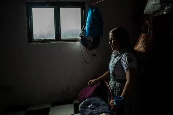 دختر نوجوانی در نمایه پشت یک صندلی در یک اتاق تاریک نزدیک به پنجره ایستاده است؛ کیف‌های دستی در پشت و نزدیک به او آویزان هستند.