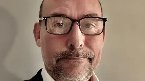 استیفن وال‌تون؛ نمای نزدیک از استیفن که موهای کوتاه قهوه‌ای، به‌تدریج از پیشانی عقب‌نشین شده، و ریش نازکی دارد؛ او عینک‌های مربعی با فریم سیاه و یک کت‌وشلوار تیره به تن دارد.