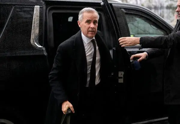 مارک کارنی از یک خودرو مشکی خارج می‌شود؛ دست شخص دیگری درهای خودرو را باز نگه داشته است. او کت مشکی، پیراهن سفید و کراوات تیره به تن دارد.