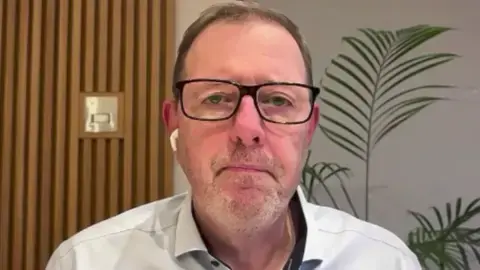 مارک مک‌کال موهای کوتاه، نازک و قهوه‌ای روشن دارد؛ عینکی با فریم مشکی به چشم دارد و پیراهنی روشن پوشیده است.