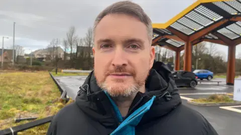 شین هاجن در فضای باز ایستاده است؛ یک کت مشکی و آبی به تن دارد و در پس‌زمینه خودروها دیده می‌شوند.