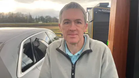 جیسون در کنار خودروی خود ایستاده است؛ یک پلیس خاکستری به تن دارد.