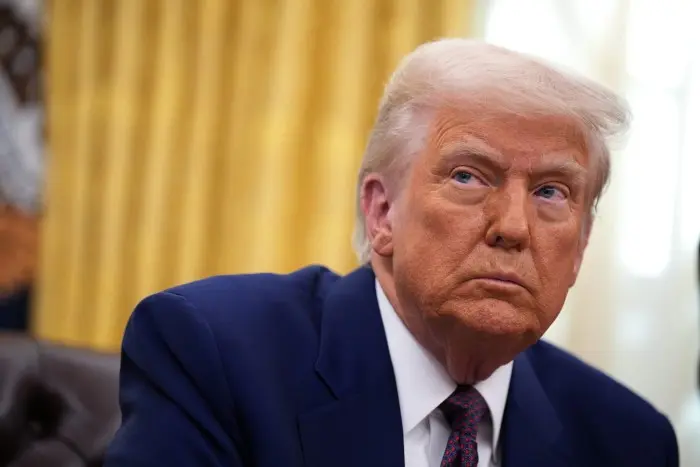 دونالد ترامپ به سمت کنار نگاه میکند در حالی که در دفتر بیضی نشسته و کت تیره و کراواتی با طرح قرمز به تن دارد.