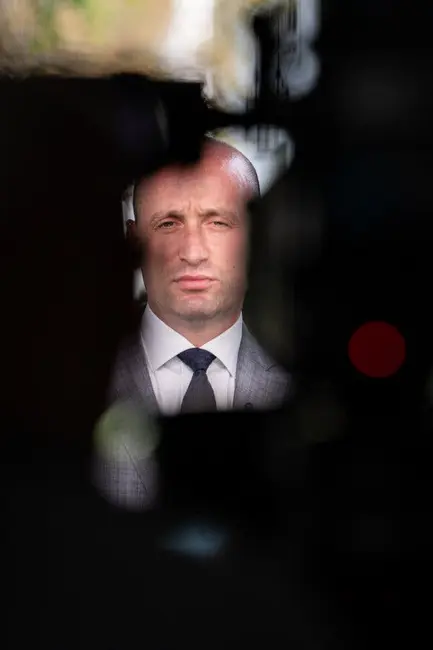 استفن میلر در حین یک مصاحبه تلویزیونی، پشت تجهیزات فیلمبرداری که به صورت سایهنما دیده میشوند، قرار گرفته است. او کت و شلوار طوسی و کراوات تیره به تن دارد.