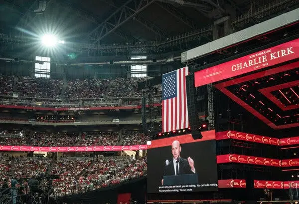 تصویر استفن میلر روی یک صفحه تلویزیون بزرگ در کنار صحنهای در یک استادیوم سرپوشیده بزرگ دیده میشود. پرچم آمریکا و پیامی با مضمون "به یاد چارلی کرک" در نزدیکی او روی صحنه نمایش داده شده است.