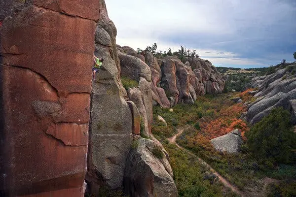 یک صخره‌نورد در حال بالا رفتن از دیواره عمودی یک صخره سرخ‌رنگ است. در دوردست، صخره‌های خاکستری‌رنگ بر فراز بوته‌زارهای سبز و نارنجی که با یک مسیر خاکی قطع شده‌اند، کشیده شده‌اند.