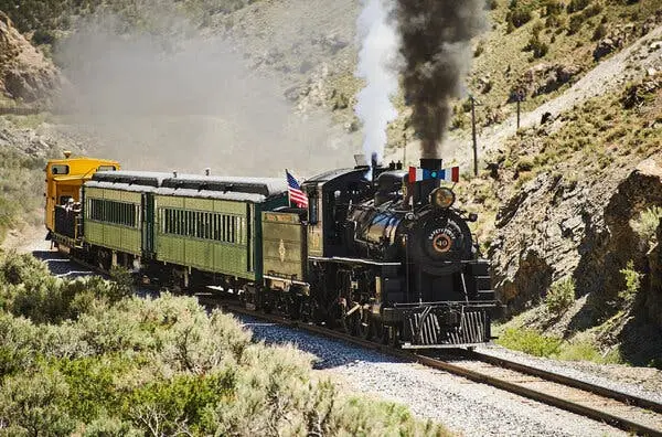 یک لوکوموتیو که از جلو دود سیاه و از پشت بخار سفید بیرون می‌دهد، سه واگن را روی ریلی در میان تپه‌های پوشیده از چمن می‌کشد.