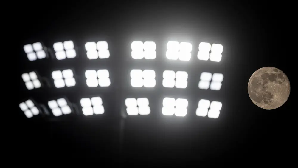 ماه کامل خاکستری در سمت راست ردیفی از چراغ‌های سیل‌زمین استادیوم در برابر آسمان شب سیاه می‌درخشد.