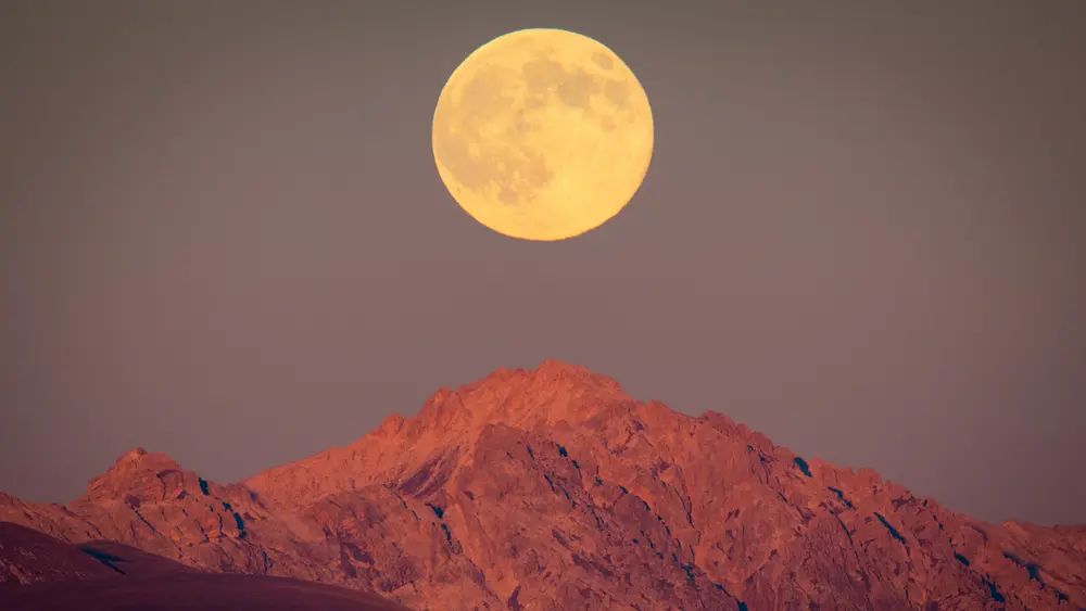 ماه کامل زردرنگ در حال طلوع بالای کوه سنگی در برابر آسمان خاکستری-آبی کم‌رنگ تصویربرداری شده است.