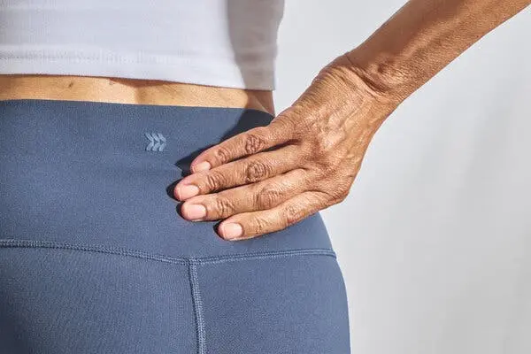 نمای نزدیک زنی که با دست خود پشت پایین خود را می‌گیرد. او یک پیراهن سفید و شلوار ورزشی آبی به تن دارد.