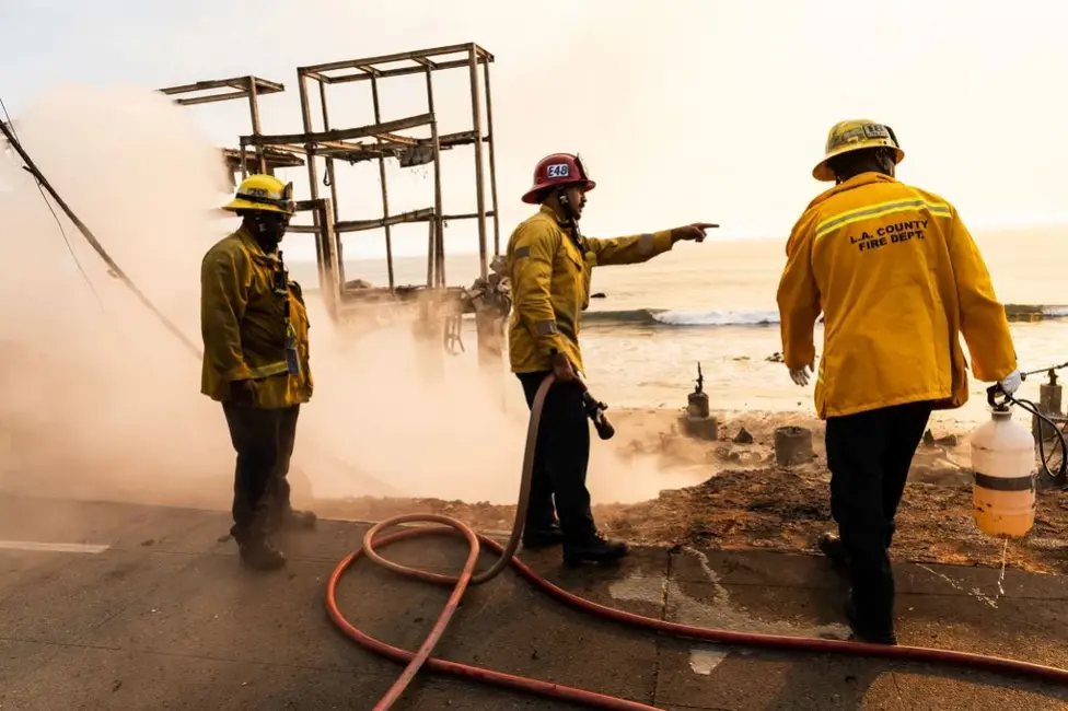 سه آتش‌نشان در نزدیکی لبهٔ خانه‌ای که پیش از این به‌دشت دریا نگاه می‌کرد و در اثر یک آتش‌سوزی جنگلی سوزانده شد، ایستاده‌اند. بخشی از ساختاری باقی‌مانده در پس‌زمینه قابل رؤیت است، در حالی که دود زیر خراله‌ها به‌بالا می‌آید.