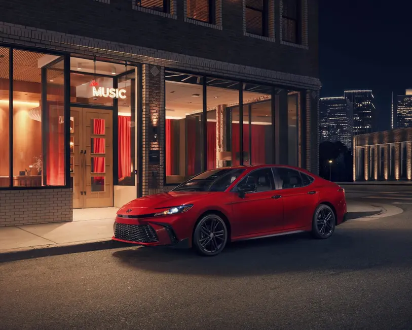 سدان قرمز پارک شده در خیابان شهری در شب، مقابل ساختمانی با پنجره‌های بزرگ و تابلو نئونی «موزیک زنده»