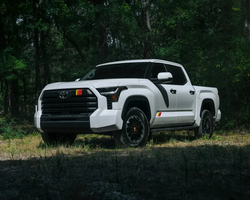 شاحنه سفید تویوتا با گریل مشکی و برچسب‌های خط قرمز-زرد، پارک شده روی چمن در دل جنگل