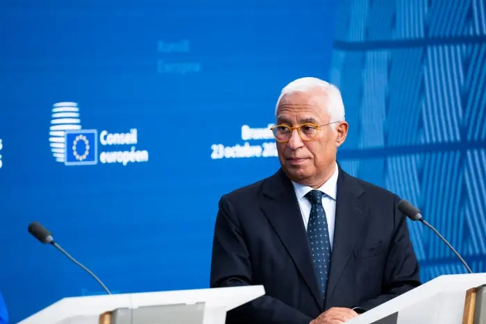 فردی با لباس رسمی، شامل عینک و کت و شلوار، در سکوی سخنرانی با پس‌زمینه آبی که لوگوهای شورای اتحادیه اروپا و متن نشان می‌دهد