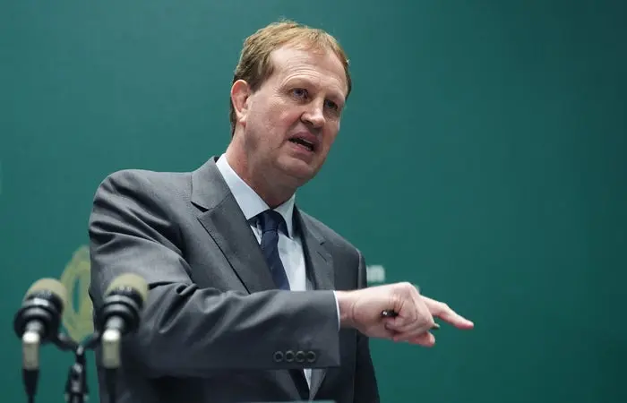 مردی در کت و شلوار با حرکات پرشور هنگام سخنرانی در مقابل میکروفون‌ها و پس‌زمینه ساده