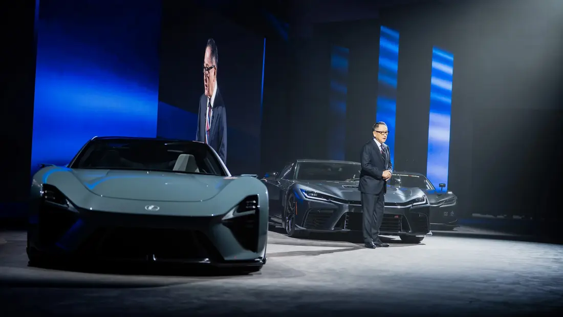 مردی در کت و شلوار ایستاده کنار دو خودروی اسپرت خاکستری تیره بر روی صحنهای با پنلهای نور عمودی آبی.