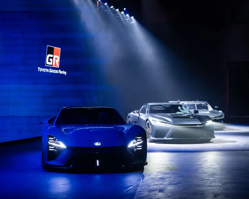 خودروی اسپرت آبی لکسس با چراغهای روشن در مقابل دو خودروی اسپرت نقرهای زیر نورهای اسنادی در یک نمایشگاه تاریک که لوگوی تویوتا گازوو ریسینگ بر روی دیوار نصب شده است.