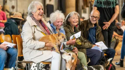 گروهی از افراد – سه زن و یک مرد – در خدمت برکت‌دادن حیوانات داخل کلیسا می‌نشینند و می‌خندند؛ سگی کوچک قهوه‌ای‑رنگ و لَکش‌لک‌شده بر روی زانوهای یکی از زنان نشسته است که توسط باسِت هاند بوییده می‌شود.